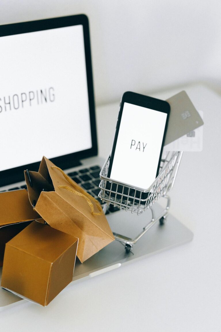 A miniature shopping cart holds a smartphone and credit card on a laptop, symbolizing online shopping.