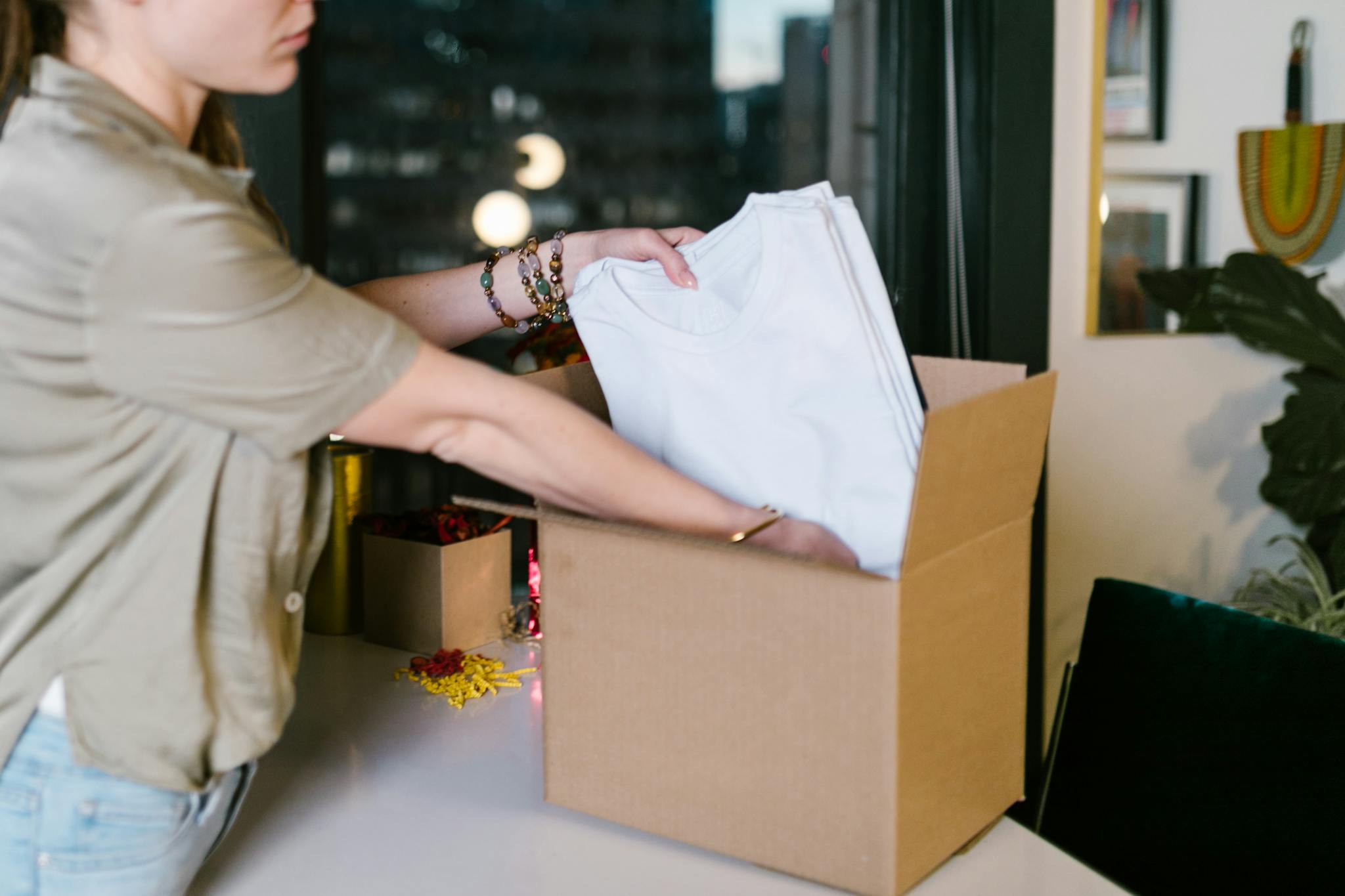 A woman packs white t-shirts into a cardboard box, set in a modern indoor environment.