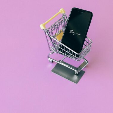 Mobile phone in mini cart on pink surface, symbolizing online shopping.